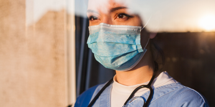 Frontline worker wearing a stethoscope and mask looks out at the sunset through a workplace window.