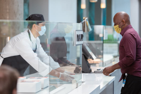 Man in mask picks up parcel at counter from retail worker in mask, apron, and hat