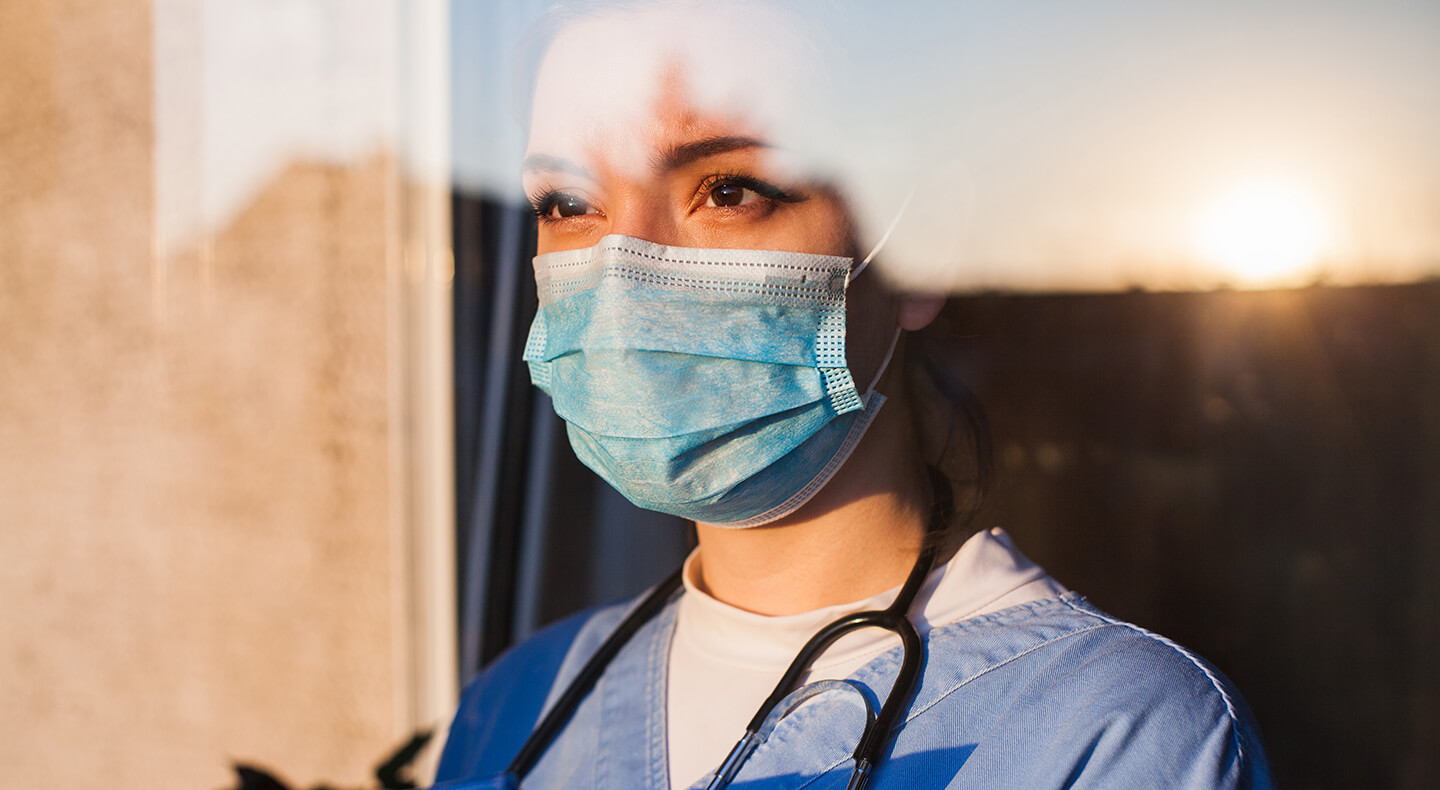 Frontline worker wearing a stethoscope and mask looks out at the sunset through a workplace window.