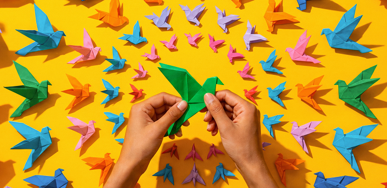Hands folding origami shapes, representing the careful shaping of new organizational structures as work is redesigned around human judgment and AI.